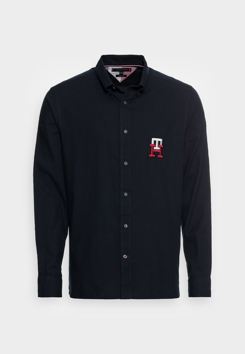 Chemise noire à manches longues avec col et boutons, avec un logo "TH" brodé en rouge et blanc sur la poitrine gauche.