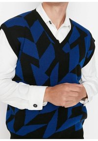 Uomo che indossa un maglione senza maniche blu e nero con motivo geometrico sopra una camicia bianca a maniche lunghe, mani incrociate davanti.