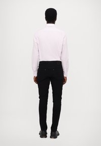 Chemise à manches longues rose clair à rayures, associée à un jean noir à coupe slim. Le modèle est de dos, mettant en valeur la tenue superposée. Chaussures noires.