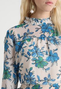 Blouse fleurie en tissu léger avec des motifs de fleurs bleues et vertes, col froncé et taille élastique.