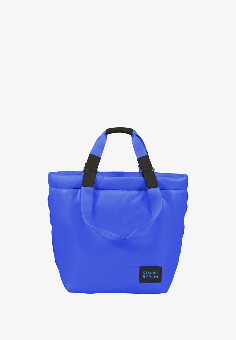 Blå vadderad axelväska med rektangulär form, med svarta detaljer, dubbla handtag upptill och en etikett på framsidan. Hållbart material.