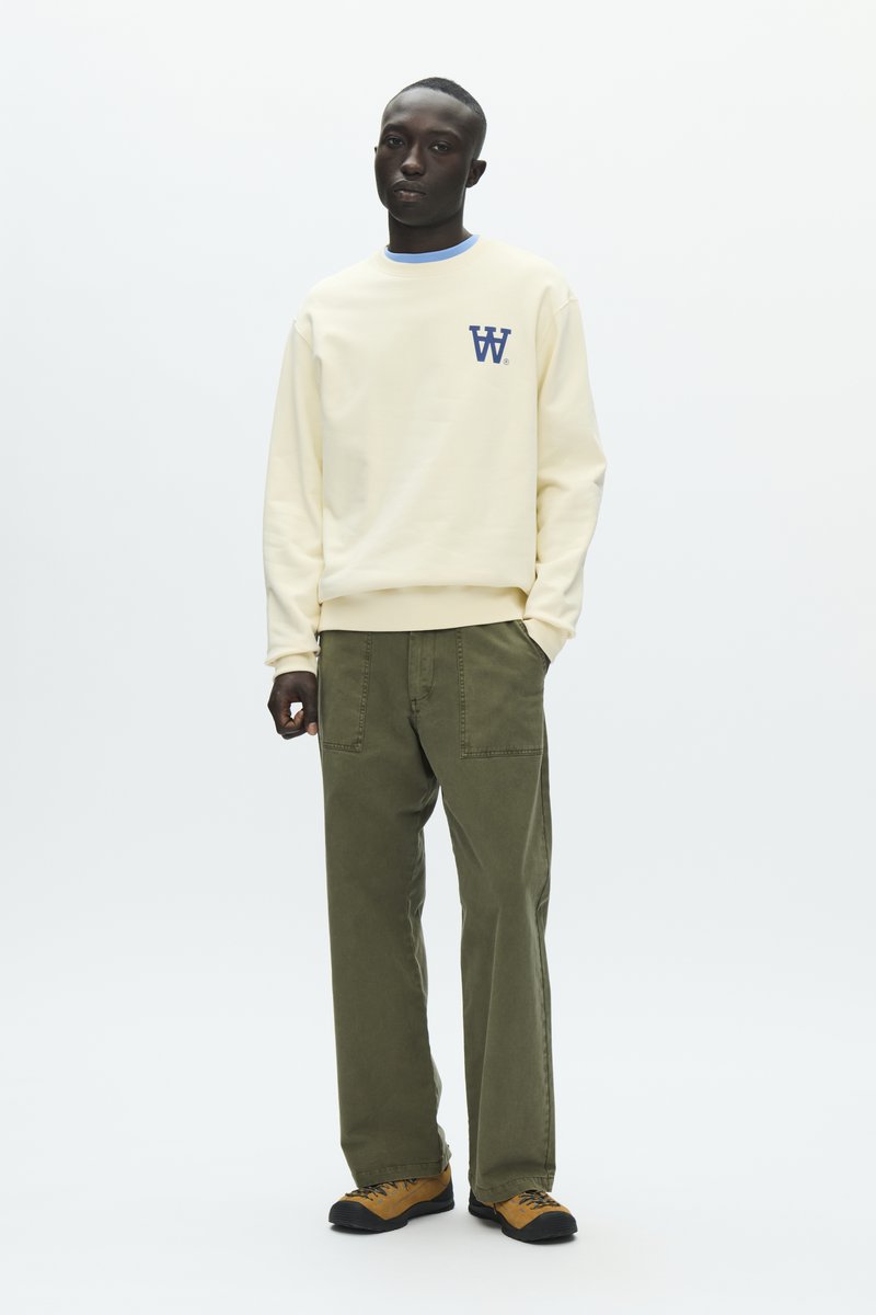 Homme debout les mains dans les poches, portant un sweat-shirt crème avec un logo bleu, un pantalon vert olive et des chaussures de randonnée marron sur fond blanc.