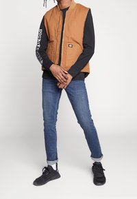 Brązowa bezrękawnik z przednim zamkiem i dwiema kieszeniami nałożona na czarną koszulę z długim rękawem, zestawiona z niebieskimi dżinsami i czarnymi trampkami.