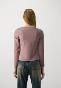 Ilgi piedurkņu tops mauve audumā, ar kvadrātu formas kakla izgriezumu un pieguļošiem siluetu. Saskaņots ar augsto jostasvietu zilajiem džinsiem ar izbalētiem akcentiem.