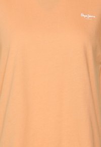 Světle oranžové tričko vyrobené z měkké tkaniny. Na levé straně má malé bílé logo. Jednoduchý, klasický design s krátkými rukávy.