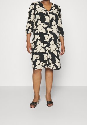 Kvinde iført en sort kjole med hvide blomsterprint, trekvartærmer, knapper foran og sorte åbne hæle.