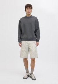 Grauer Pullover mit gerippten Bündchen, kombiniert mit beigen Cargo-Shorts, die über mehrere Taschen und einen knie-langen Schnitt verfügen.