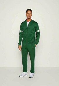 Green sports tracksuit made of smooth fabric, featuring white stripes on sleeves and pants, zip-up jacket, and fitted design, paired with white sneakers.