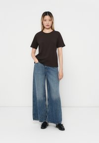 Brūns īsām piedurknēm t-krekls apvienots ar platām zila denim džinsām. Džinsiem ir gaiši izbalējusi apakšmalā. Modelis stāv, ar vienu roku kabatā.