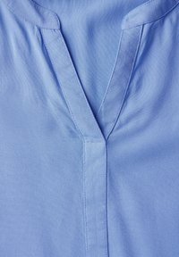 Camicia azzurra chiara con scollatura a V, texture morbida e dettagli di pieghe sottili sulla parte anteriore. Maniche di lunghezza media.