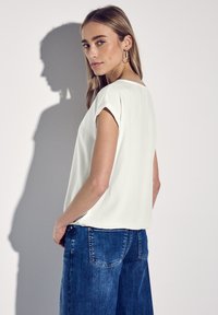 Bílý krátký rukáv top vyrobený z hladké tkaniny, v kombinaci s tmavě modrými džíny; modelka se pózuje bokem, což ukazuje siluetu a stín.