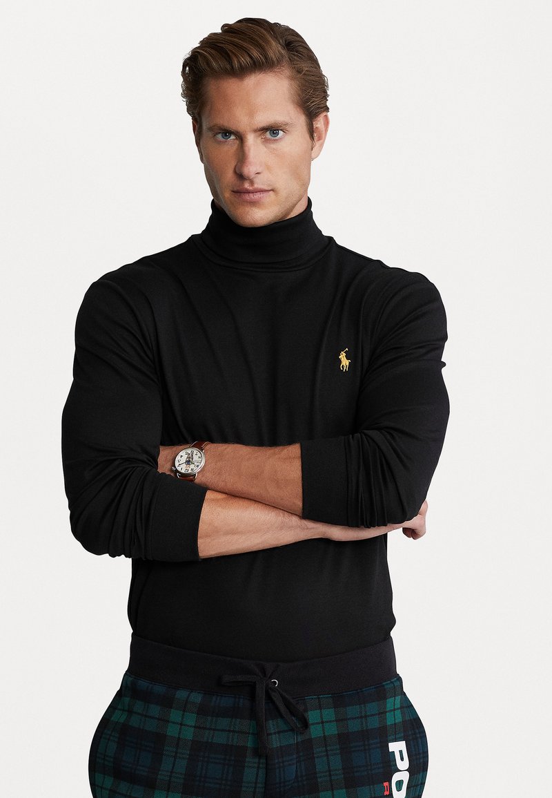 Camiseta de cuello alto negra con un diseño ajustado, combinada con pantalones de chándal a cuadros en verde y azul marino. Presenta un logo dorado en el pecho y un reloj en la muñeca.