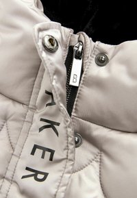 Beige quiltet jakke med høj krave, der har en sølvlynlås og sort foring. Et logo er synligt nær lynlåsen. Der er sølvknapper til stede.