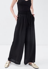 Pantalon noir à jambes larges avec une taille élastique et un cordon de serrage ; tissu léger ; poches latérales ; texture lisse ; associé à des chaussures dorées à enfiler.