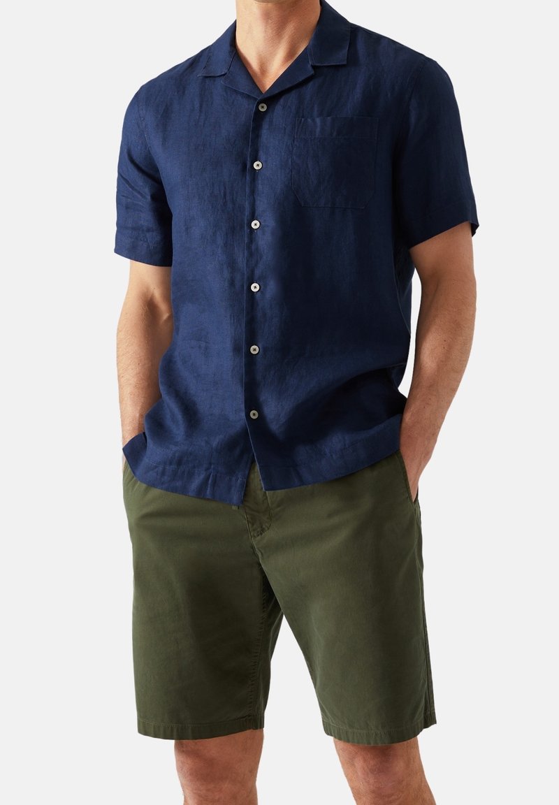 Mann trägt ein marineblaues kurzärmliges Hemd mit Knöpfen und olivgrüne Shorts, die Hände in den Taschen, steht vor einem einfarbigen Hintergrund.