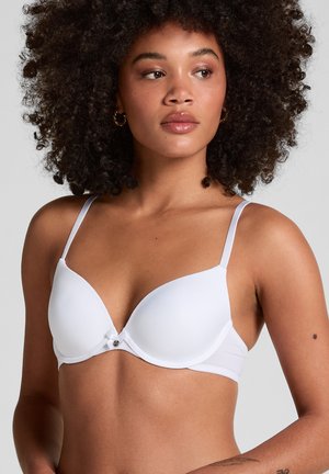 Femme aux cheveux bouclés portant un soutien-gorge blanc à bonnets moulés avec des bretelles fines et un petit nœud, regardant légèrement vers sa gauche.