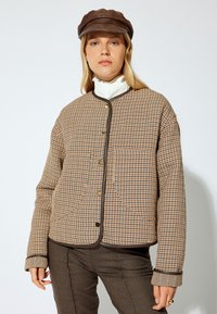 Vadderad jacka i brun och beige rutigt mönster med rund krage, framknappar, två stora fickor och kontrasterande detaljer.