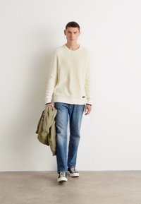 Biały sweter w prążki, niebieskie dżinsy, czarno-białe sneakersy. Model trzyma zieloną kurtkę, stojąc na tle białego, gładkiego tła.