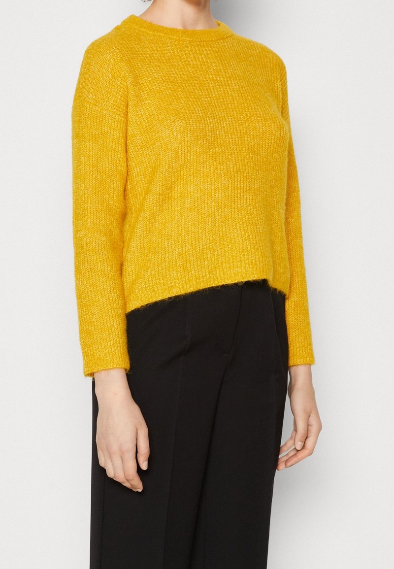 Gul stickad tröja med långa ärmar, rundad halsringning och kortad nederkant. Texturerad tyg ger djup. Bärs med svarta byxor.