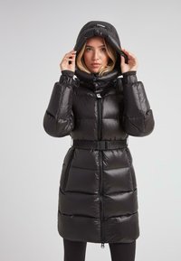 Mujer ajustando la capucha de un abrigo de plumas negro con cinturón, con cremallera frontal y bolsillos con cremallera, de pie contra un fondo claro y liso.