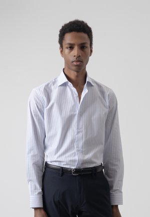 Joven con camisa de vestir blanca de manga larga con rayas, metida en pantalones negros con cinturón negro, de pie frente a un fondo liso.