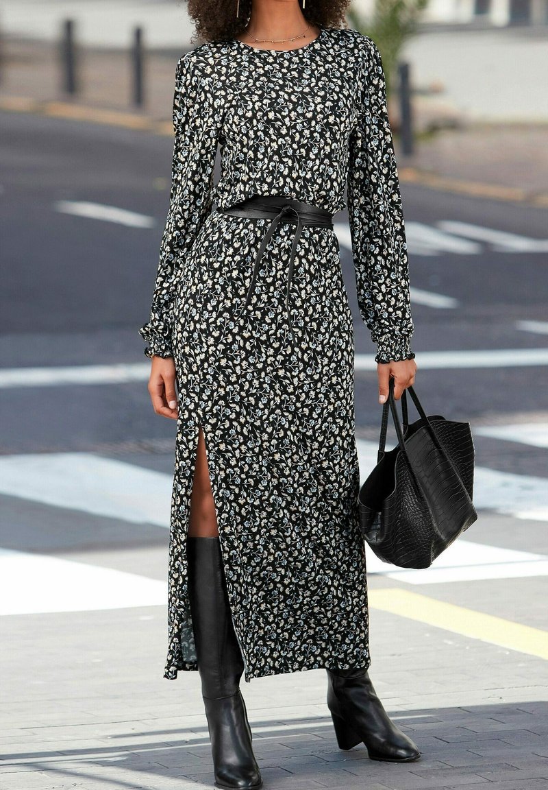 Femme portant une longue robe noire à fleurs avec une fente latérale, une ceinture noire, des bottes noires montant jusqu'au genou et tenant un grand sac à main noir dans une rue en ville.