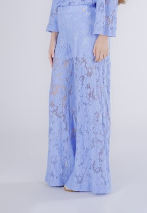 Žena oblečená ve vysokým pasem, širokými nohavicemi světle modrých krajkových kalhot s květinovým vzorem a odpovídajícím krajkovým topem, stojící na jednobarevném pozadí.