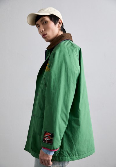 Grøn jakke med en brun krave, der har en patch på ærmet og synlige knapper. Stoffet ser glat ud med en afslappet stil.