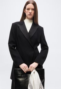 Γυναίκα με ίσια καστανοκίτρινα μαλλιά, που φοράει μαύρο σακάκι και λευκό τοπ, κρατώντας μια μαύρη δερμάτινη τσάντα με ένα λευκό φουλάρι με σχέδιο.