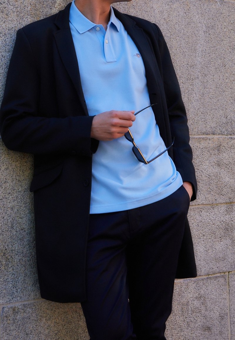 Homme appuyé contre un mur en pierre, portant un manteau noir, un polo bleu clair, un pantalon foncé, tenant des lunettes de soleil noires dans la main droite.