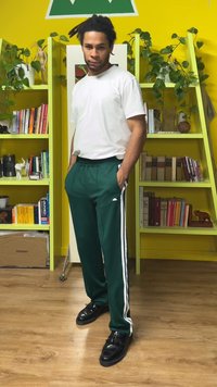 Grønne treningsbukser med hvite sidestriper, kombinert med en enkel hvit t-skjorte, brukt med svarte sko. Innendørs omgivelser med gul bakgrunn.