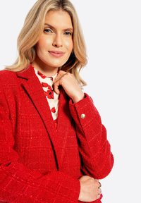 Blazer rosso con texture, colletto a bavero, caratterizzato da un motivo a quadri e dettagli con bottoni dorati. Indossato sopra una blusa con stampa floreale.