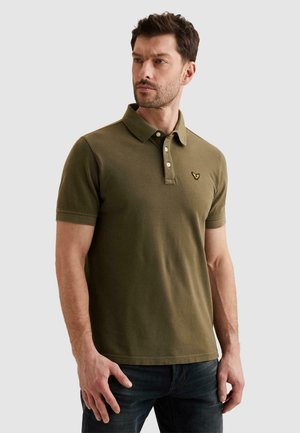 Hombre con cabello corto y castaño, vistiendo una camiseta tipo polo de manga corta color verde oliva y jeans oscuros, de pie frente a un fondo claro y liso.