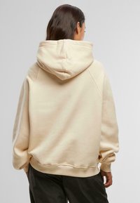 Beige színű, oversized kapucnis pulóver, húzózsinórral, bordázott mandzsettákkal és aljjal, sima pamut textúrával. Ráépített ujjak a laza illeszkedés érdekében.