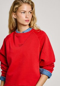 Felpa rossa con maniche raglan, logo ricamato sul petto e colletto di camicia in denim blu che spunta da sotto. Texture morbida.