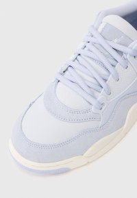 Primo piano di una sneaker blu pallido con pannelli in suede e mesh, lacci bianchi e suola color crema.