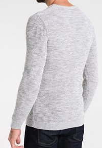 Maglione grigio lavorato a maglia con tessuto a rilievo, scollo tondo e bordi a coste su polsini e orlo, indossato sopra jeans scuri.