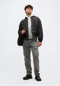 Νεαρός άνδρας που φοράει μαύρο μπουφάν bomber, γκρι φούτερ, γκρι παντελόνι cargo, μαύρα αθλητικά παπούτσια, κρατώντας μαύρο σακίδιο πλάτης, στέκεται μπροστά σε απλό φόντο.