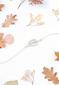 Silverarmband med ett bladformat hänge som har intrikata detaljer och klara stenar, omgiven av torkade löv på en vit bakgrund.
