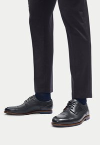 Černé kožené společenské boty se šněrováním, kulatou špičkou, kontrastním hnědým prošíváním a nízkým podpatkem, v kombinaci s tmavými kalhotami a ponožkami.