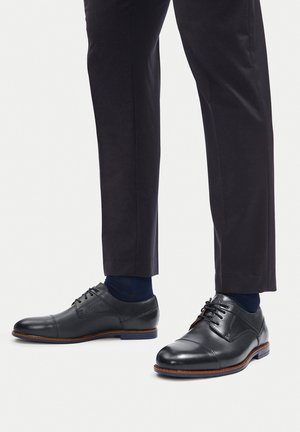 Čierne kožené spoločenské topánky s šnurovaním, okrúhlou špičkou, kontrastným hnedým prešívaním a nízkym podpätkom, spárované s tmavými nohavicami a ponožkami.