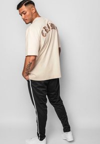Beige oversized t-shirt med brun kursiv skrift på ryggen, parret med sorte joggers og hvide sneakers, der viser en afslappet pasform.