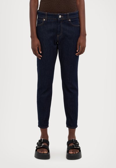 Σκούρο μπλε τζιν παντελόνι με στενή εφαρμογή, κοντό μήκος, πέντε τσέπες και καφέ ραφή, συνδυασμένο με μαύρες πλατφόρμες.