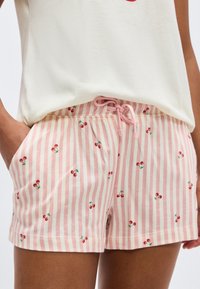 Γυναίκα που φοράει ροζ και λευκά κάθετα ριγέ σορτς με μικρά τυπώματα κερασιών και ροζ κορδόνι, συνδυασμένα με λευκό τοπ.