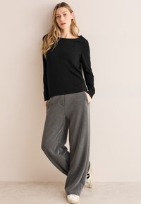 Maglietta nera a maniche lunghe con scollo ampio, abbinata a pantaloni della tuta grigi larghi, dotati di tasche laterali e di un tessuto testurizzato. Sneakers bianche.