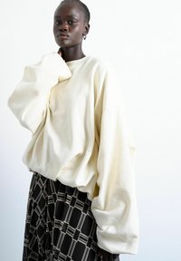 Personne à la peau foncée portant un sweat-shirt surdimensionné blanc cassé avec des manches volumineuses et une jupe noire à motifs, posant devant un fond blanc uni.