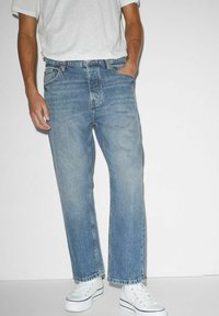 Man med ljusblå raka jeans, vit t-shirt och vita platåskor, som står mot en enkel vit bakgrund.