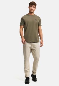 Olivgrünes T-Shirt mit kurzen Ärmeln und Rundhalsausschnitt, ausgestattet mit einem kleinen schwarzen Logo, kombiniert mit hellbeigen Hosen und schwarzen Schuhen.