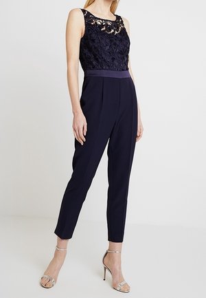 Mørkeblå jumpsuit med blomstret blondetop, ærmeløs design, tætsiddende talje og tilspidsede ben. Har en ensfarvet taljebånd og skræddersyet silhuet.
