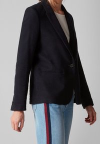 Blazer marine en tissu doux avec un bouton unique, revers crantés et poches latérales, associé à un jean bleu clair avec des rayures rouges.